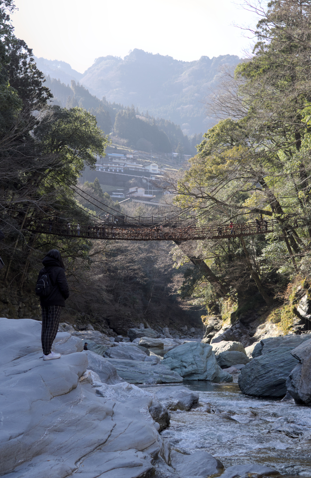 View of a valley from its bottom, rocks at its base with a person standing on them and a suspension bridge in the distance.